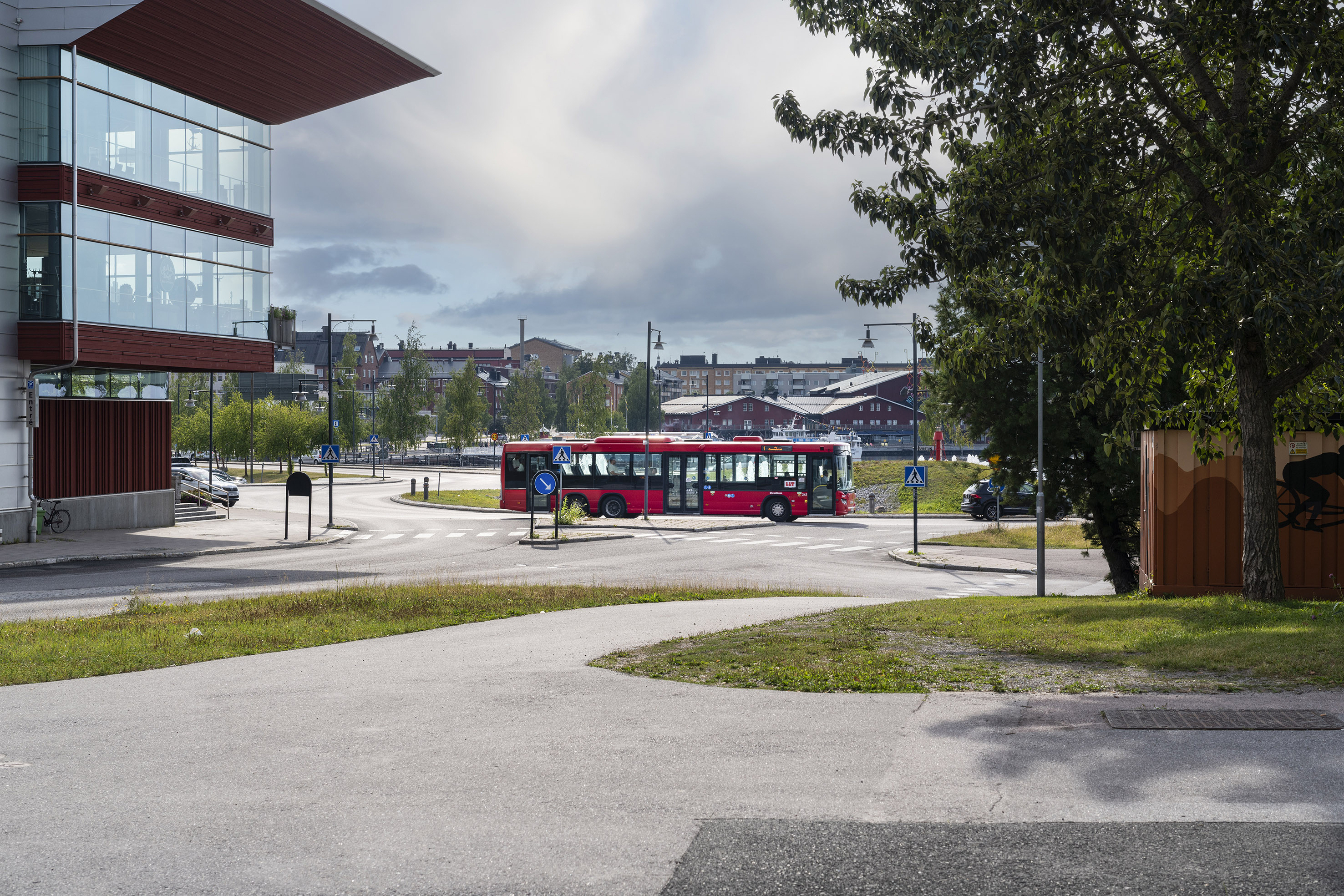 Röd stadstrafikbuss på en gata med korsning i en stadsområde, omgiven av moderna byggnader och träd.