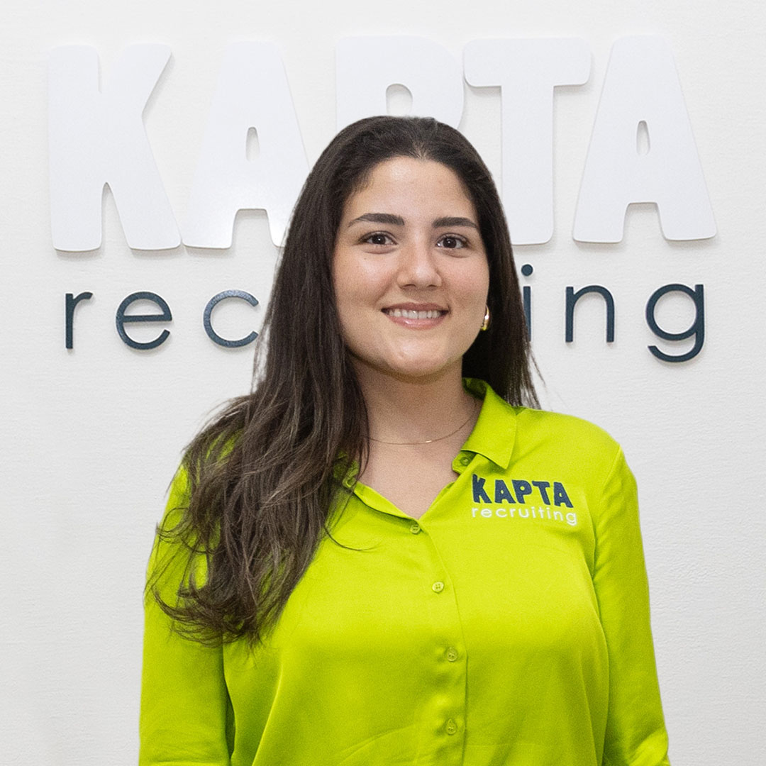 Vero, Marketing Managerin bei Kapta Recruiting steht in grüner Bluse mit KAPTA-Logo, lächelnd vor dem Schriftzug ‚KAPTA recruiting‘ an einer weißen Wand.