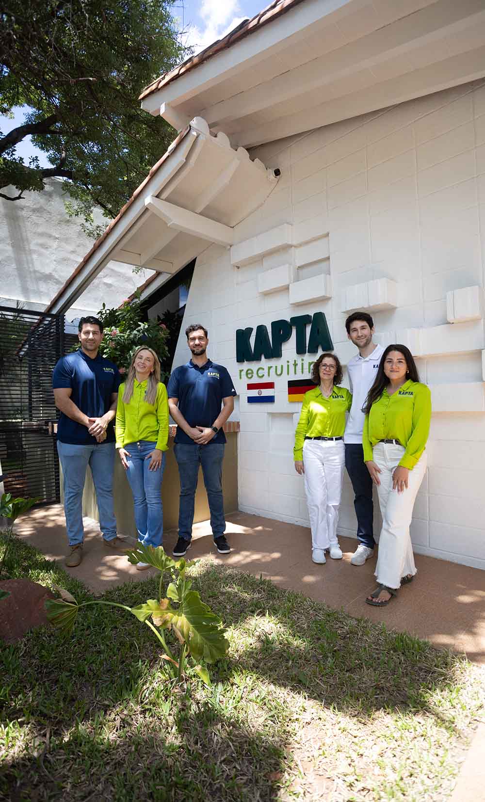 Sechs Mitarbeitende stehen lächelnd vor dem Gebäude von „Kapta Recruiting“. Auf der Wand hinter ihnen ist das Unternehmenslogo sowie die Flaggen von Paraguay und Deutschland zu sehen. Das Team trägt einheitliche Arbeitskleidung in Blau, Weiß und Grün.