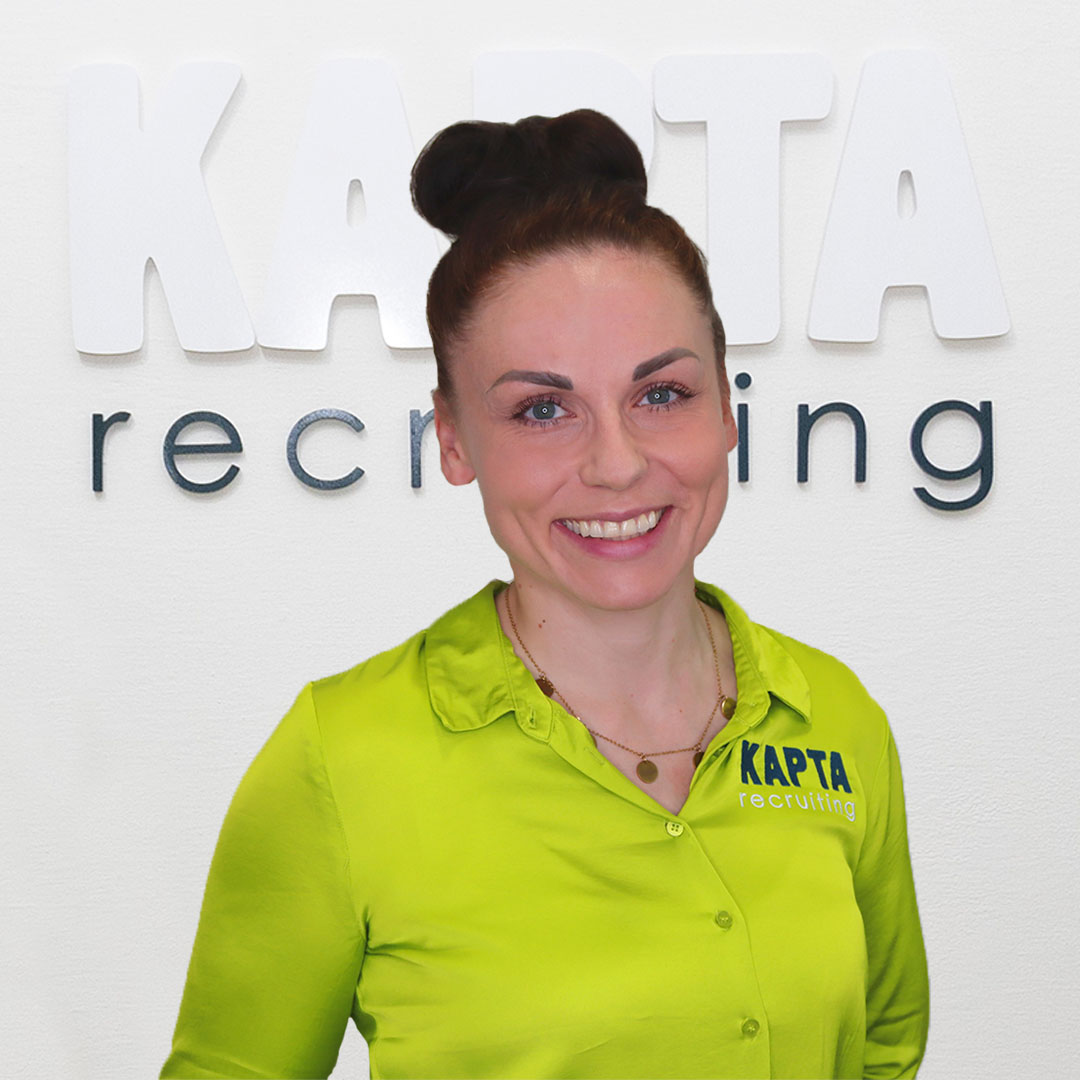 Vero, Marketing Managerin bei Kapta Recruiting steht in grüner Bluse mit KAPTA-Logo, lächelnd vor dem Schriftzug ‚KAPTA recruiting‘ an einer weißen Wand.
