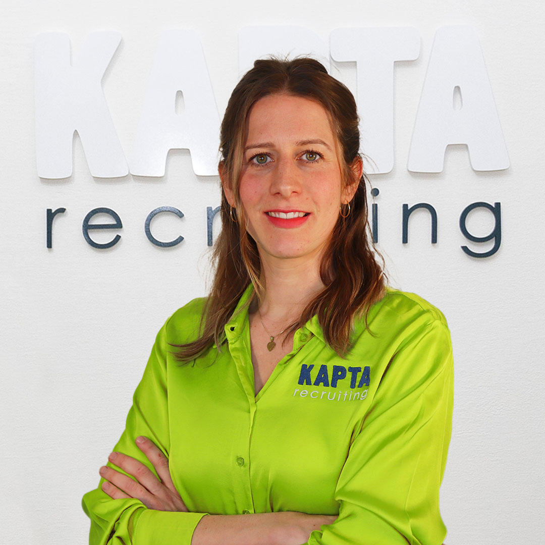 Kim, Account Managerin bei Kapta Recruiting steht in grüner Bluse mit KAPTA-Logo, lächelnd vor dem Schriftzug ‚KAPTA recruiting‘ an einer weißen Wand.