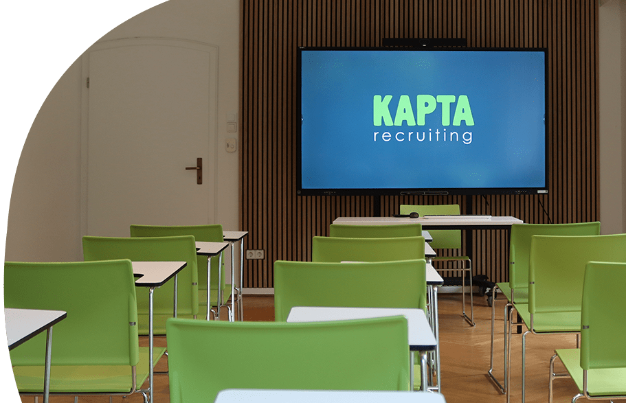 Vier Personen sitzen in einem modernen Konferenzraum um einen runden Tisch und führen ein Gespräch. Zwei Frauen tragen grüne Hemden mit dem Logo von KAPTA Recruiting, während die anderen in Weiß gekleidet sind. Auf dem Tisch liegen Mappen und Laptops, an der Wand ist das KAPTA-Logo sichtbar.