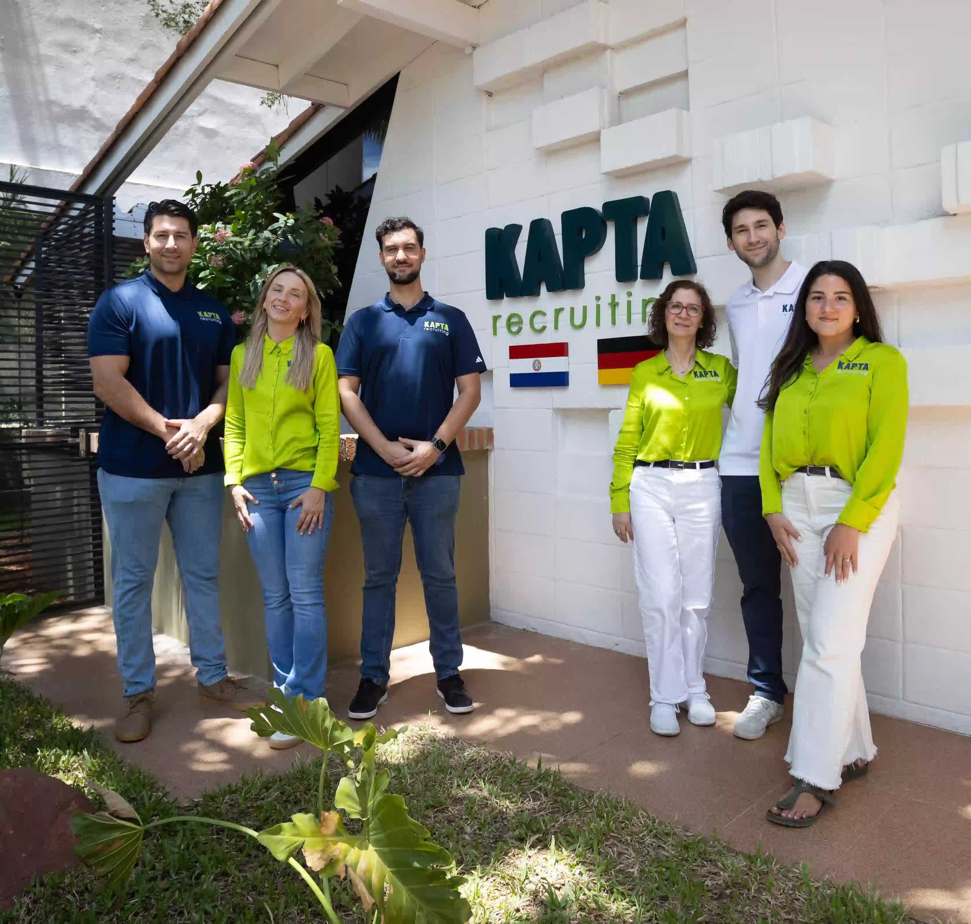 Sechs Mitarbeitende stehen lächelnd vor dem Gebäude von „Kapta Recruiting“. Auf der Wand hinter ihnen ist das Unternehmenslogo sowie die Flaggen von Paraguay und Deutschland zu sehen. Das Team trägt einheitliche Arbeitskleidung in Blau, Weiß und Grün.