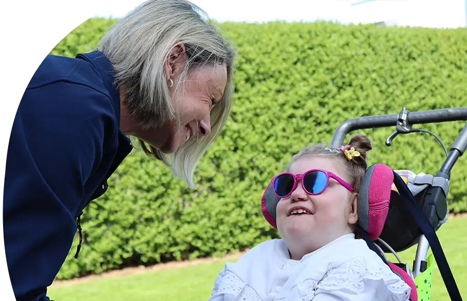 Eine lächelnde Frau mit blonden Haaren beugt sich freundlich zu einem fröhlichen Mädchen im Rollstuhl, das eine pinke Sonnenbrille trägt. Die Szene spielt sich draußen im Grünen ab und strahlt Wärme und Verbundenheit aus.