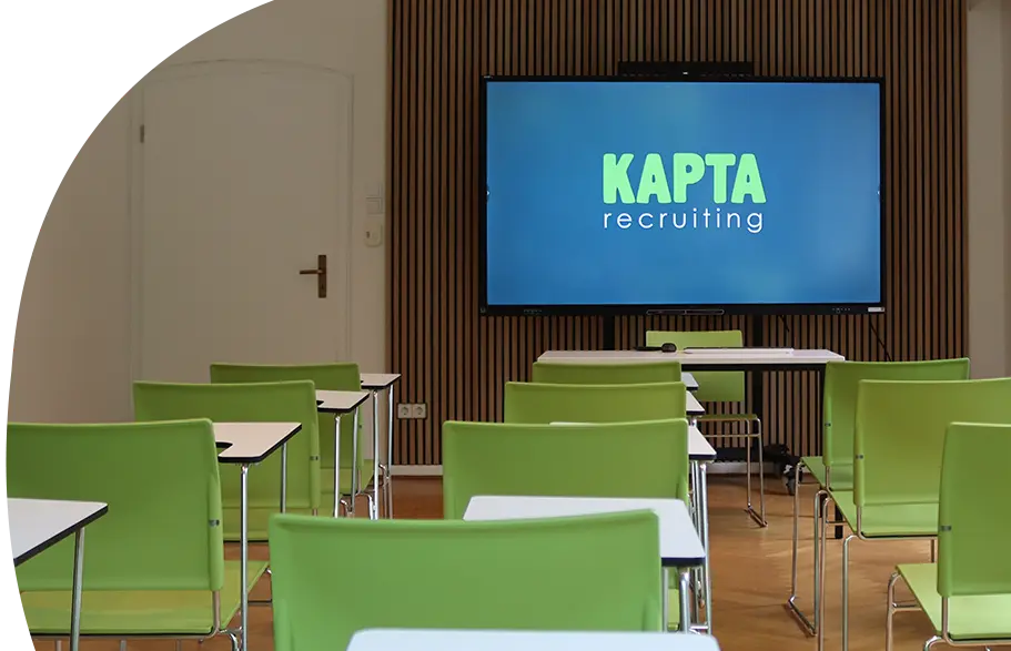 Vier Personen sitzen in einem modernen Konferenzraum um einen runden Tisch und führen ein Gespräch. Zwei Frauen tragen grüne Hemden mit dem Logo von KAPTA Recruiting, während die anderen in Weiß gekleidet sind. Auf dem Tisch liegen Mappen und Laptops, an der Wand ist das KAPTA-Logo sichtbar.