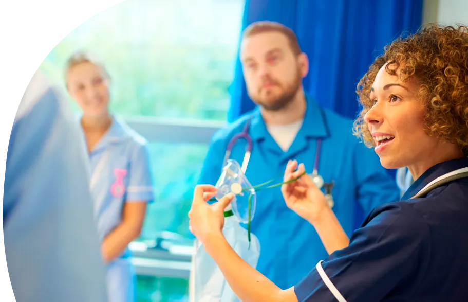 Eine Frau mit langen blonden Haaren und auffällig grünem Hemd steht mit dem Rücken zur Kamera und hält vor einer Gruppe junger Erwachsener einen Vortrag. Die Teilnehmenden sitzen in einem hellen Raum mit großen Fenstern, tragen weiße T-Shirts und wirken aufmerksam und interessiert.