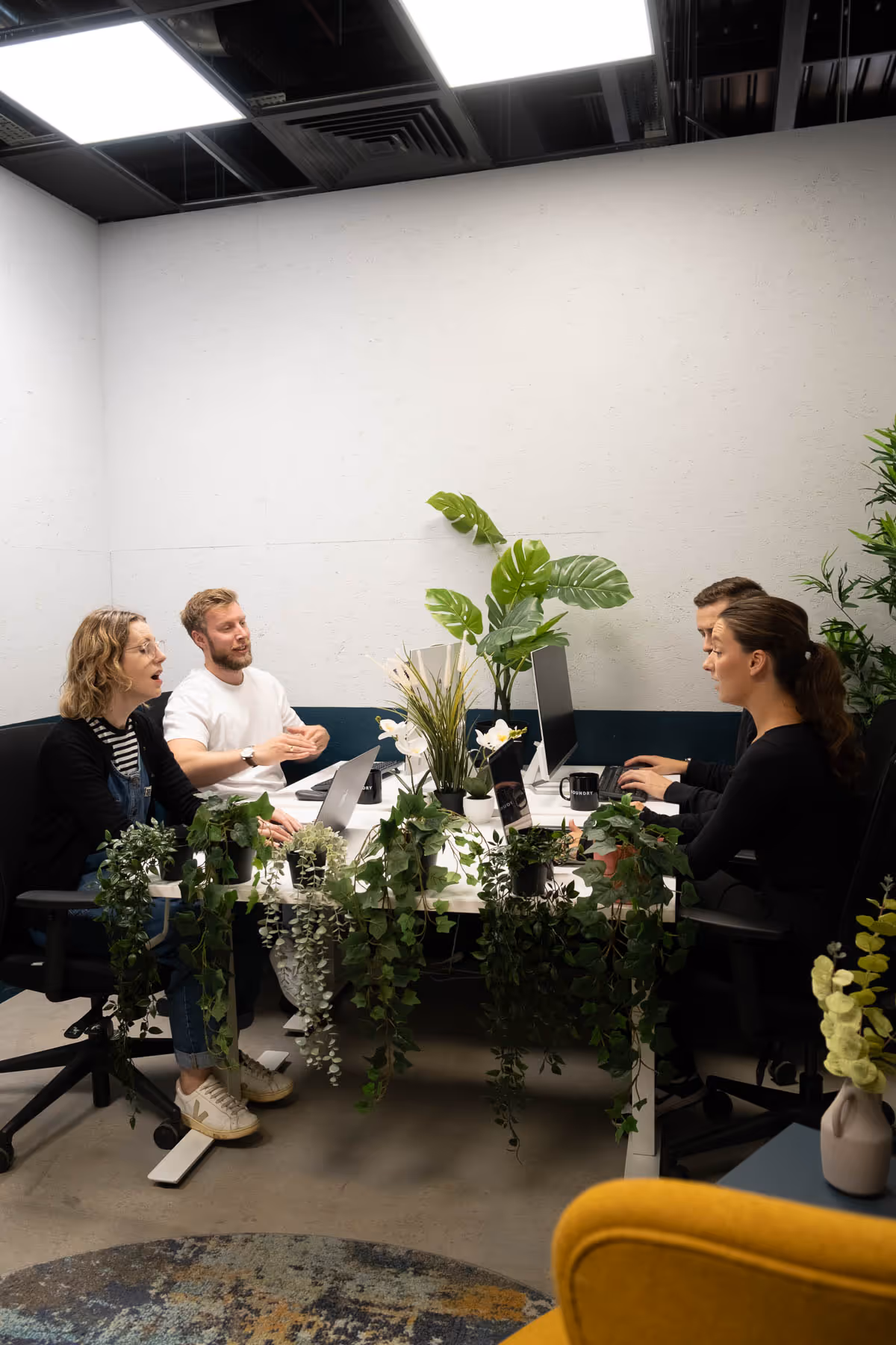 4 people at at a shared desk talking and working on laptops