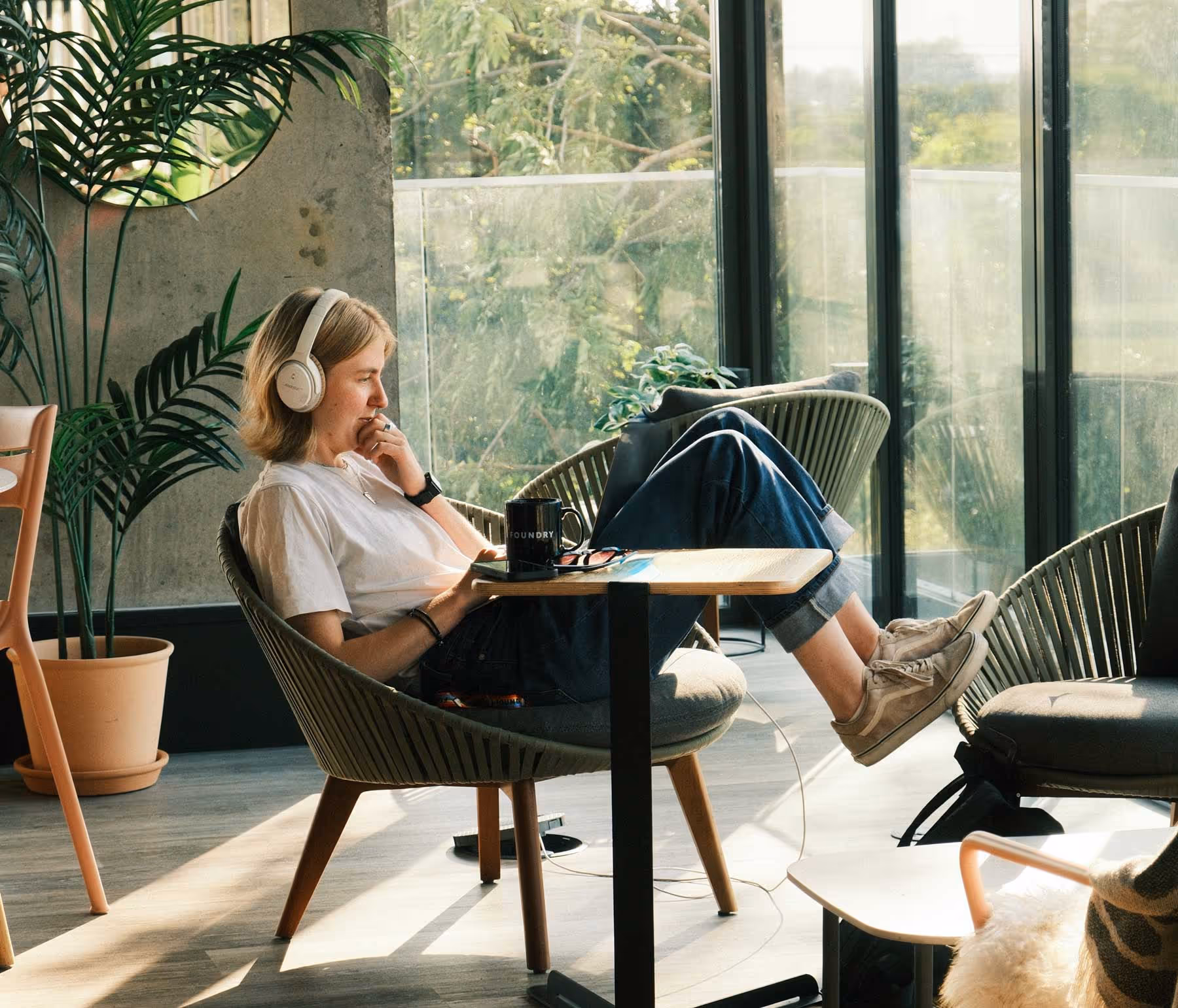 A girl sat with her feet up on a chair working on a laptop with headphones on.