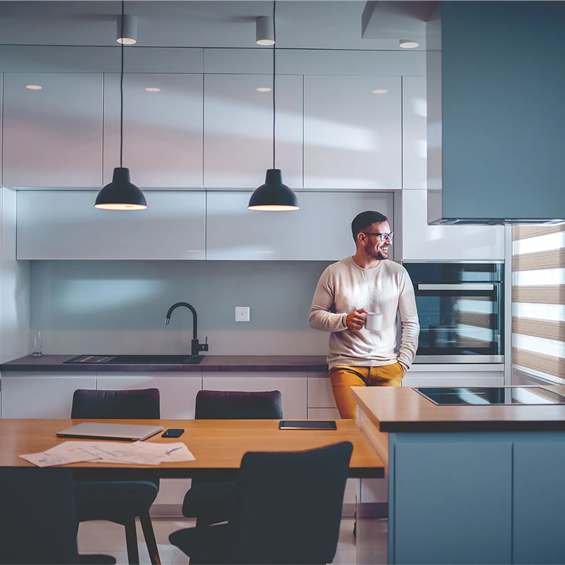 Uomo con occhiali e pantaloni color senape appoggiato a un bancone della cucina con in mano una tazza bianca, cucina moderna con armadi bianchi, rubinetto lavabo nero, due lampade a sospensione, tavolo da pranzo in legno con documenti, un laptop e uno smartphone e una finestra con tende orizzontali.