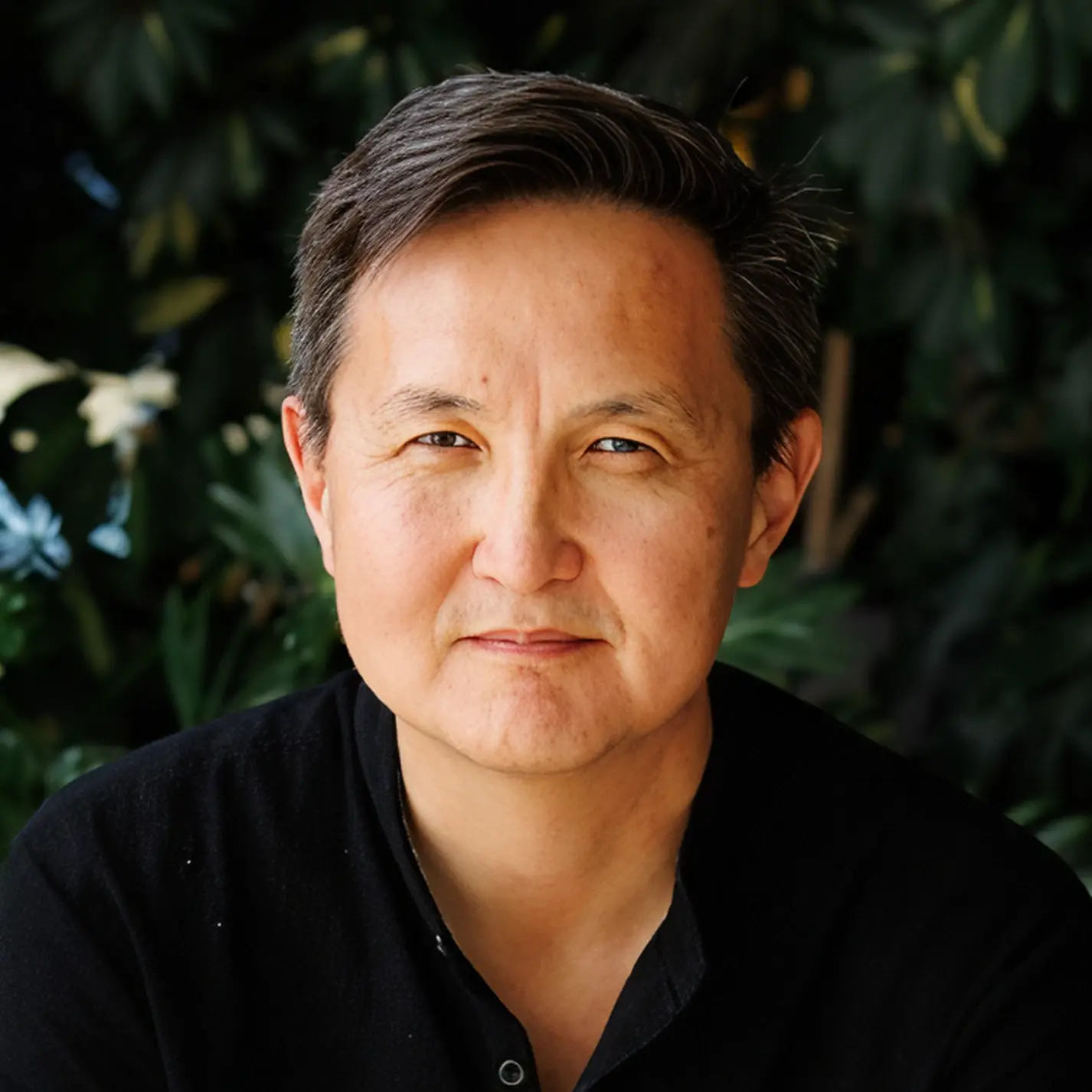Arsen Tomski. Portrait of a middle-aged man with short black and gray hair wearing a black shirt in front of leafy green plants.