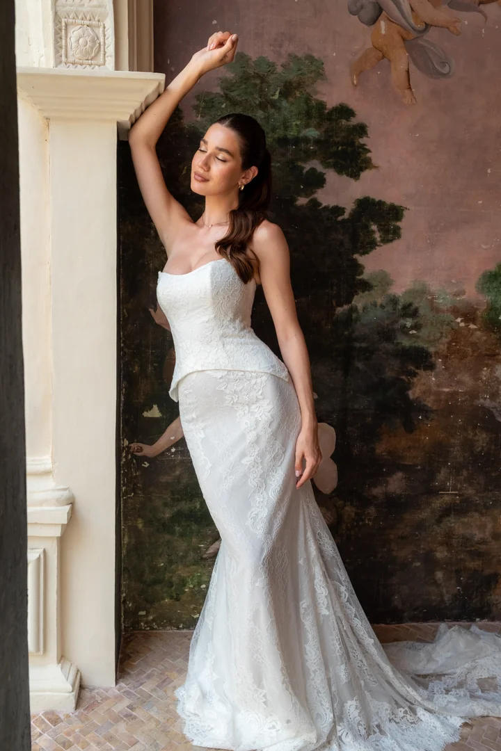 Modern romance meets timeless craftsmanship in Sorelle, a beautifully coordinated two-piece Chantilly lace bridal set. Crafted in delicate ivory lace, the structured corset bodice features a contemporary cat- eye neckline that softly frames the décolletage. The elongated bodice ﬂows into an inverted basque waistline, sculpting the waist and creating a striking, architectural silhouette that feels both reﬁned and fashion-forward. At the back, a sleek zipper closure ensures a secure and seamless ﬁt.
The matching Chantilly lace skirt hugs the ﬁgure before gently ﬂaring into an elegant train, creating a ﬂattering ﬁt-and-ﬂare silhouette that moves with effortless grace. Designed with versatility in mind, Sorelle allows the bride to transform her look throughout the day — pair the corset bodice with the Sorelle Mini Skirt for a playful second look, perfect for your ﬁrst dance or after-party celebration.