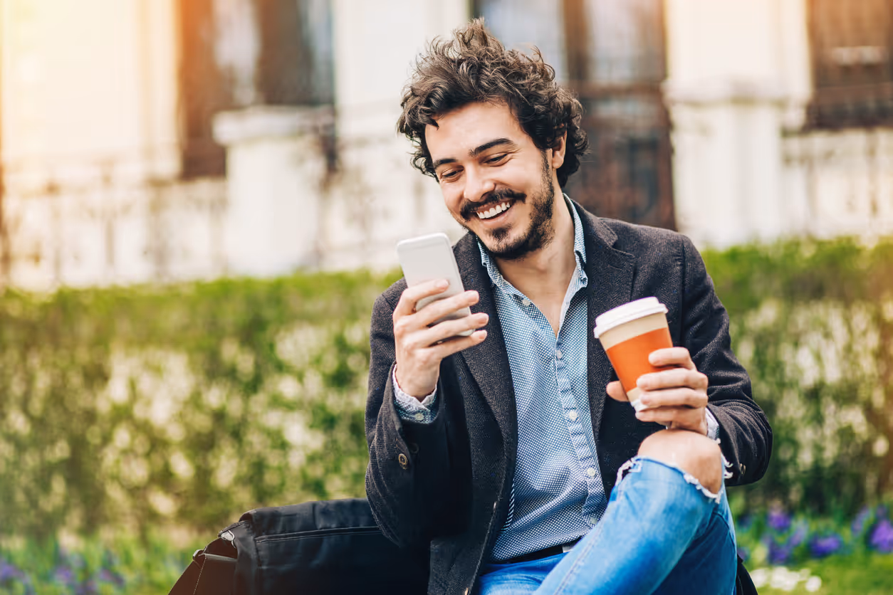 Ein Mann sitzt auf einer Bank im Freien, hält einen Kaffeebecher in der einen Hand und schaut lächelnd auf sein Smartphone