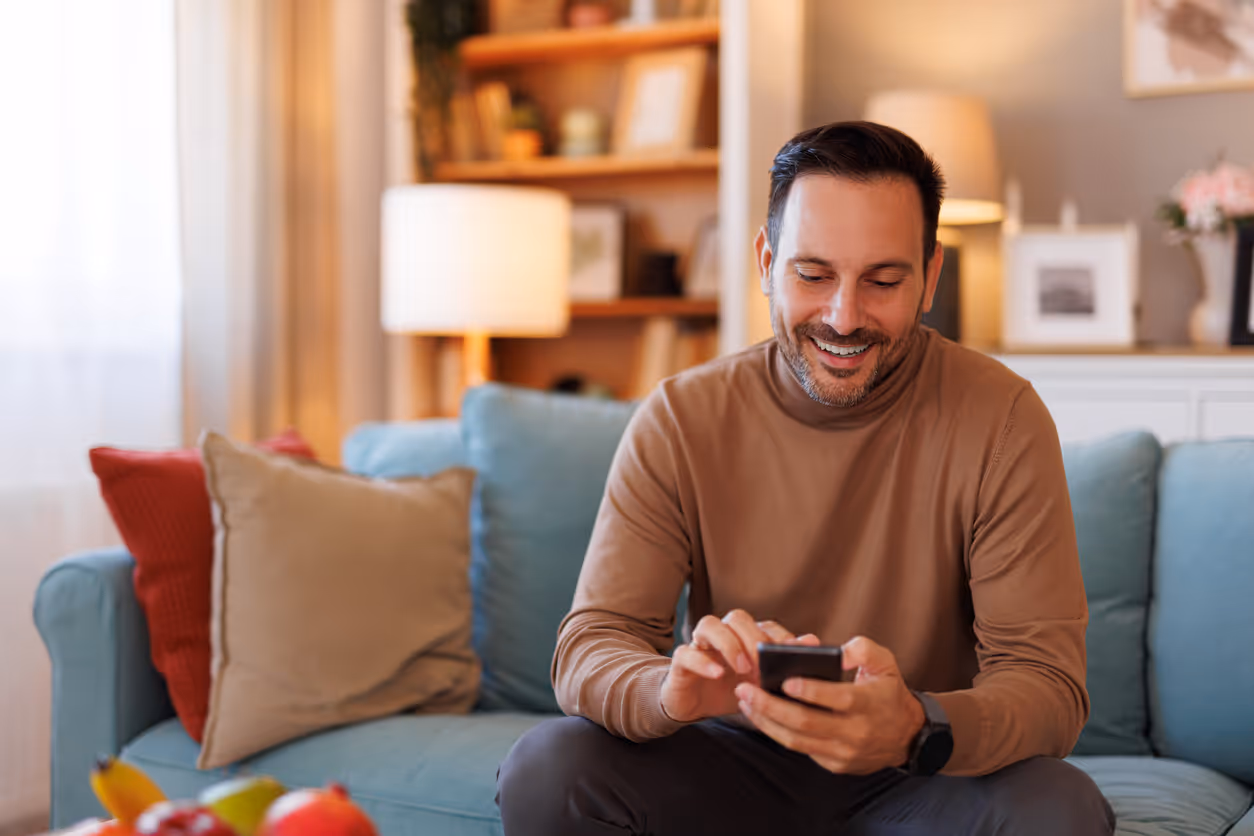 Ein Mann sitzt auf einem Sofa und lächelt, während er auf seinem Smartphone tippt