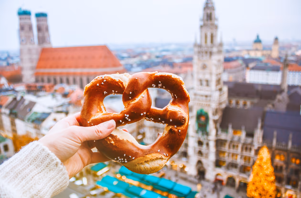 Hand mit Brezel vor dem Hintergrund des Panoramas des Münchner Platzes am Rathaus zur Zeit des Weihnachtsmarkts
