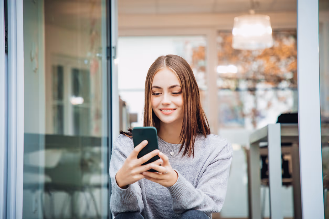 Frau, die an einem offenen Fenster steht und lächelnd auf ihr Handy blickt