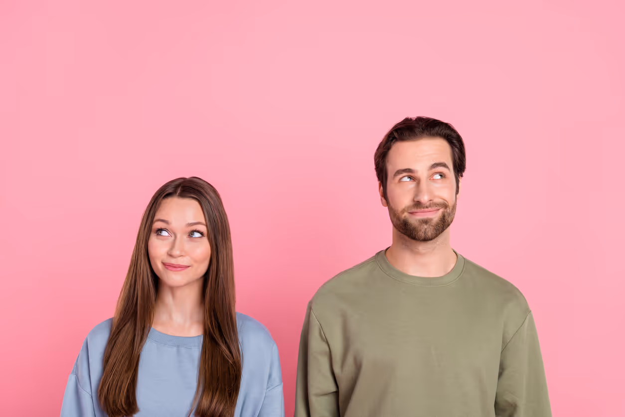 Eine Frau und ein Mann stehen vor einem pinken Hintergrund und schauen schmunzelnd in unterschiedliche Richtungen.