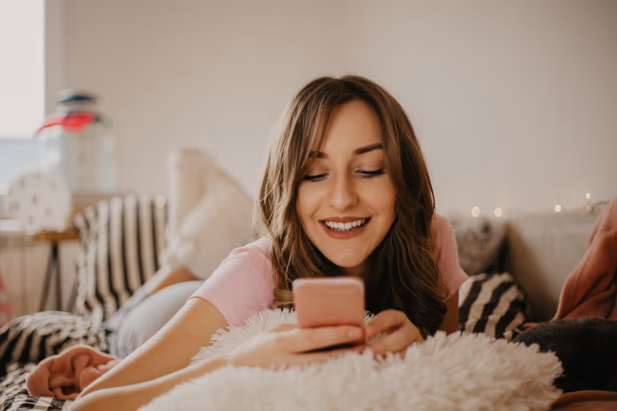 Eine Frau liegt auf dem Bauch auf einem Sofa und schaut lächelnd ihr Handy an.