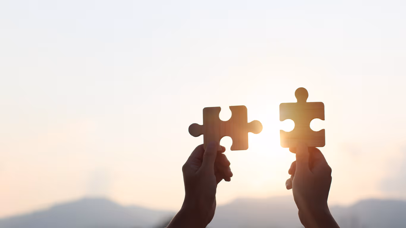 Zwei Hände, die zusammengehörende Puzzleteile vor einem Sonnenuntergang halten.