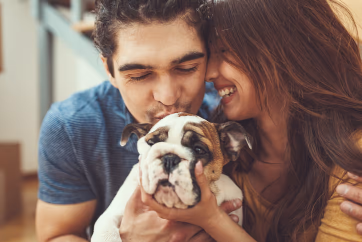 Ein junge Frau lacht. Sie wird von ihrem Partner umarmt. Beide halten gemeinsam einen Hund zwischen sich
