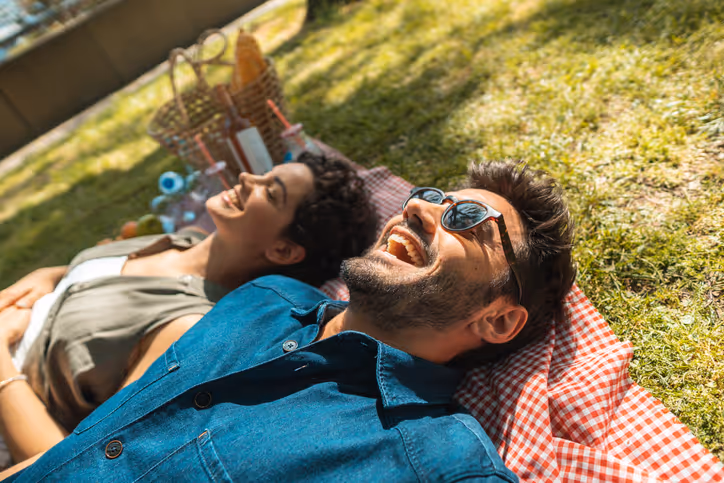 Ein Paar liegt auf einer Picknickdecke im Park, lacht und genießt den sonnigen Tag.