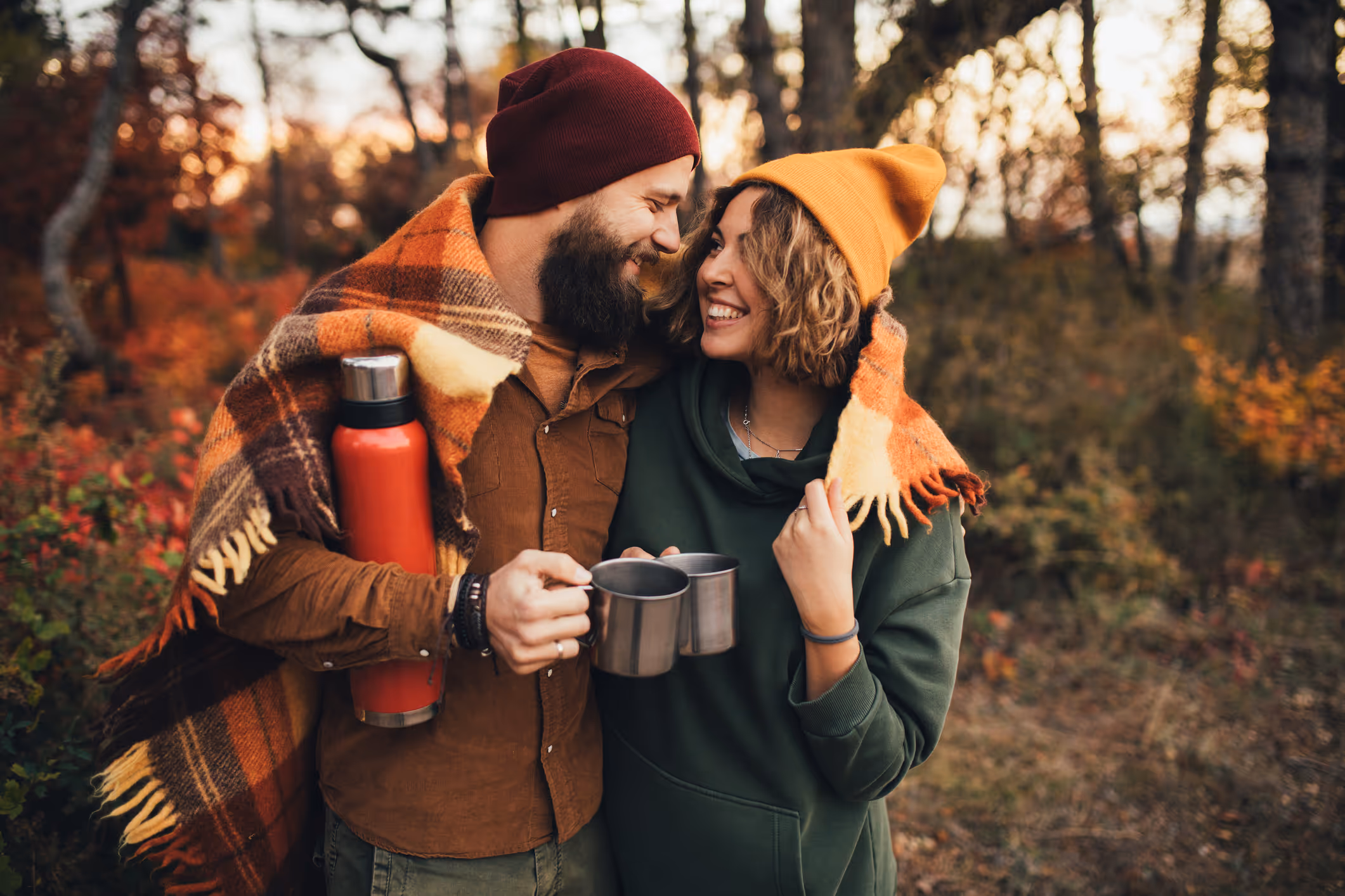 Ein Paar in warmer Kleidung umarmt sich im Wald und hält Tassen, während sie einander anlächeln.