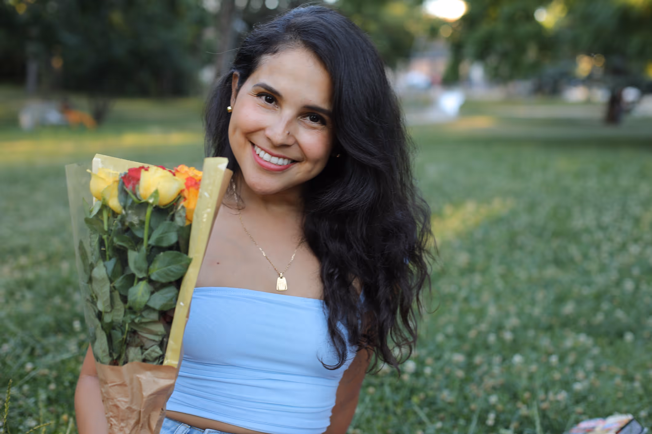 Eine Frau lächelt, hält einen Blumenstrauß mit roten und gelben Rosen und sitzt im Grünen.