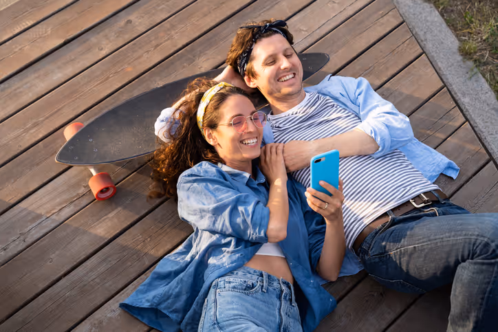 Zwei Personen liegen entspannt auf einem Holzsteg und schauen gemeinsam auf ein Smartphone