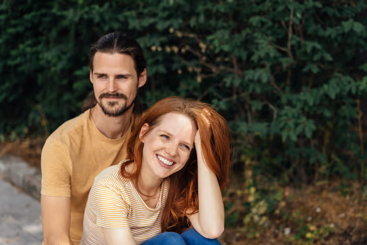 Ein lachendes Paar sitzt zusammen im Freien, umgeben von grünen Büschen und Bäumen