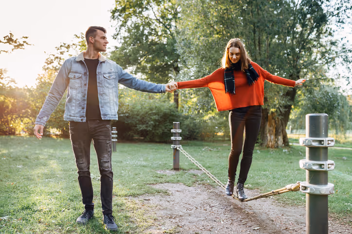  Eine junge Frau steht im Freien auf einem Balance-Seil. Ein Mann hält ihre Hand