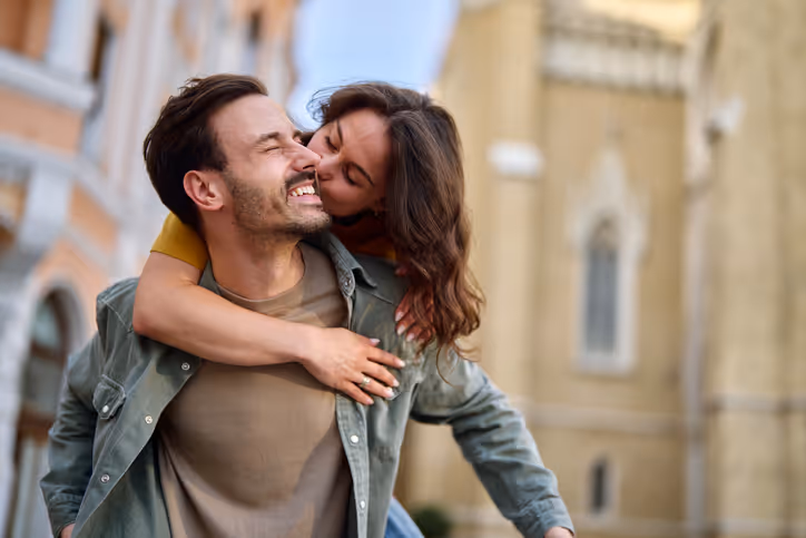 Frau wird huckepack von ihrem Freund durch die Stadt getragen und küsst ihn auf die Wange