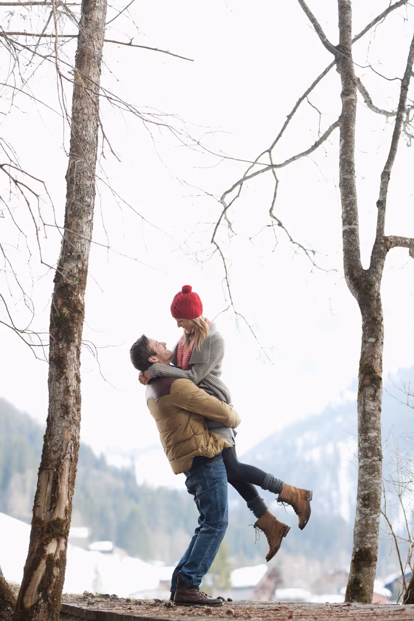 Mann hebt Frau im Schnee auf einem Waldweg mit Blick auf verschneite Berge