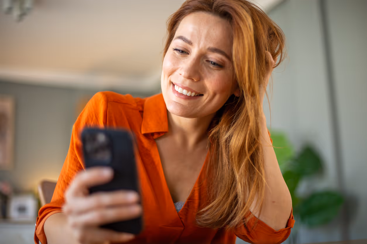 Frau mit oranger Bluse lächelt, während sie auf ihr Smartphone schaut und mit der Hand durch ihr Haar fährt