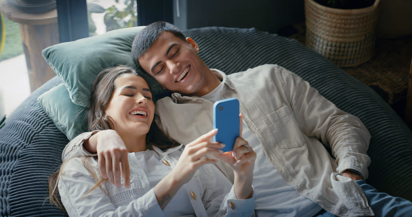 Paar liegt auf einem Sofa im Wohnzimmer und schaut zusammen auf ein Smartphone.