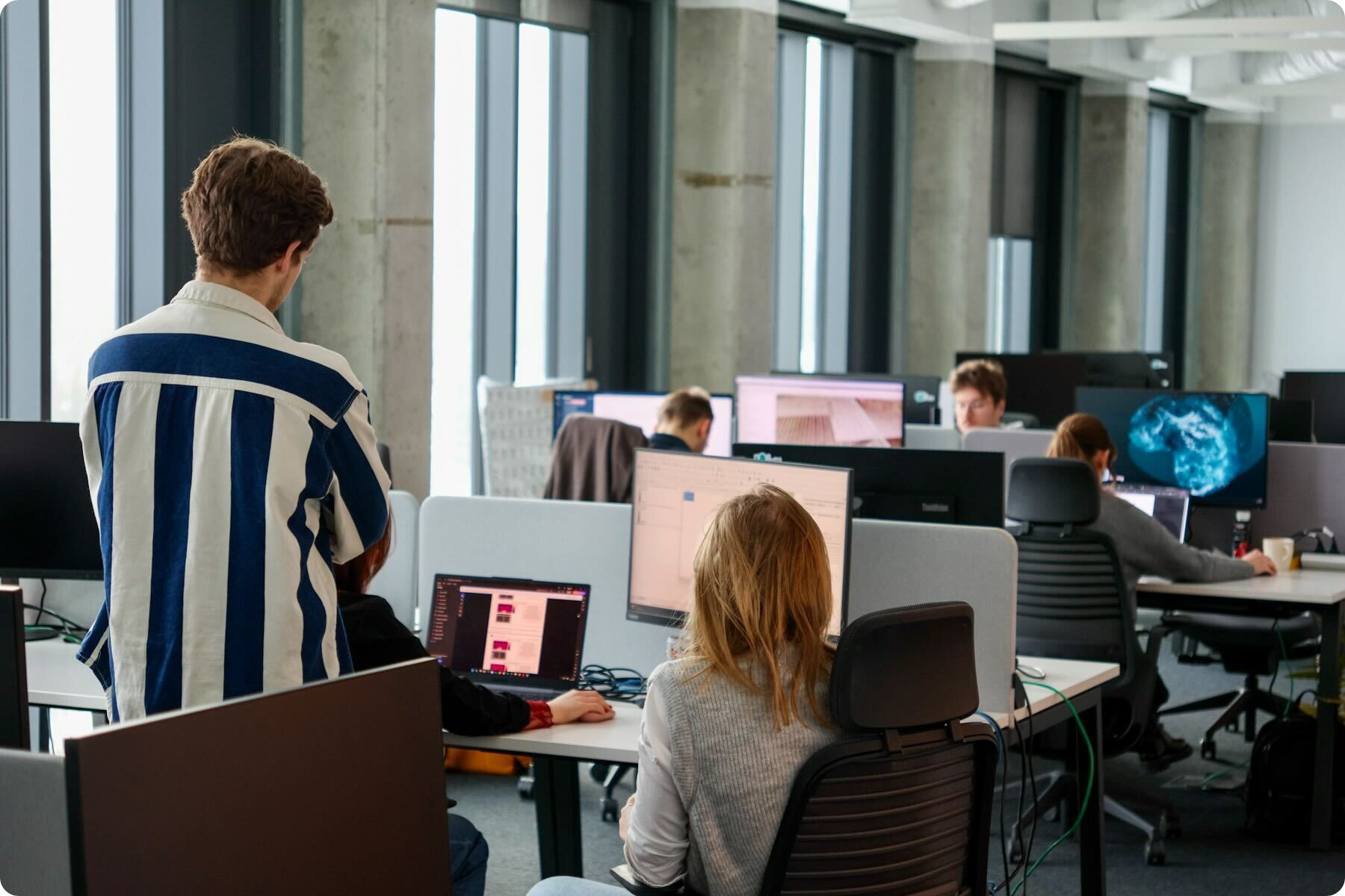 AI Clearing construction-tech team at computer workstations in a modern open-plan office, monitoring construction site progress with drone imagery, 3D terrain models, earthworks (cut/fill) analytics, QA/verification checklists, dashboards, and project reporting.