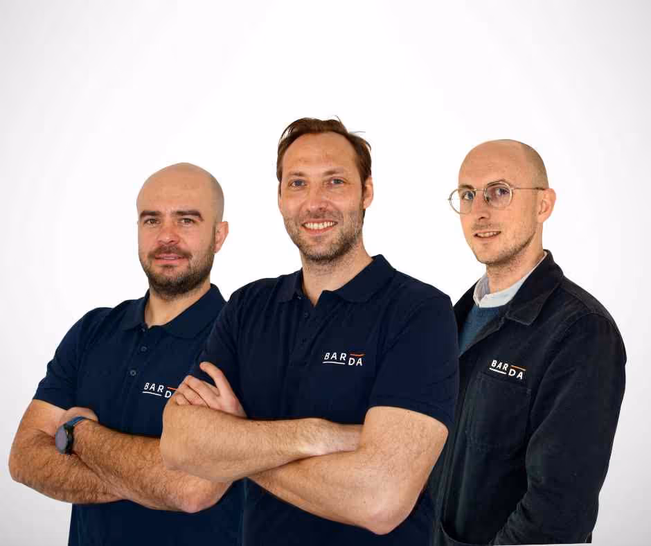 Trois hommes souriants portant des vêtements avec le logo BARDA sur fond blanc, posant avec les bras croisés.