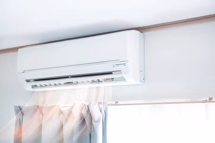 A wall-mounted air conditioner emits cool air into a sunlit room, above light gray curtains.