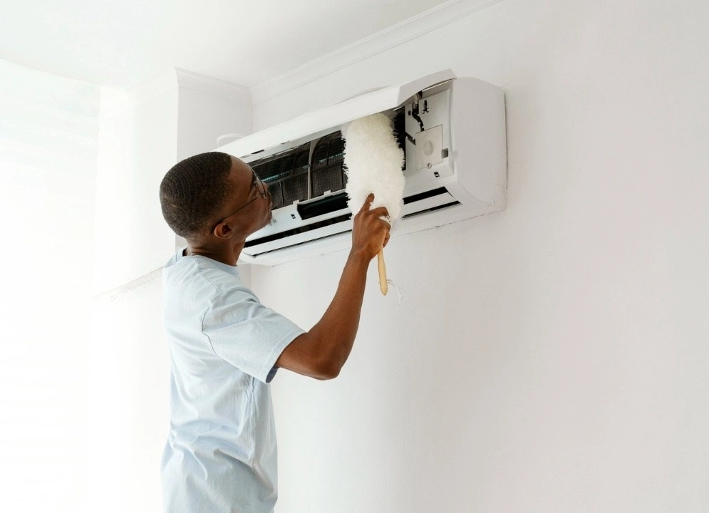A man in a light blue shirt uses a soft white duster to clean the fins and interior of a wall-mounted mini-split indoor unit.