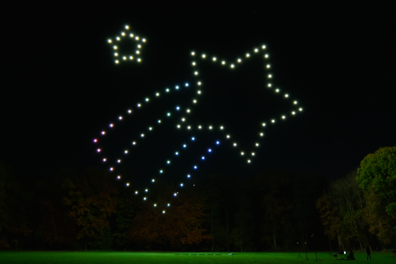 Helle Lichtpunkte formen am Nachthimmel zwei Sterne, einer größer mit Schweif, über einem dunklen Wald und grünem Feld.