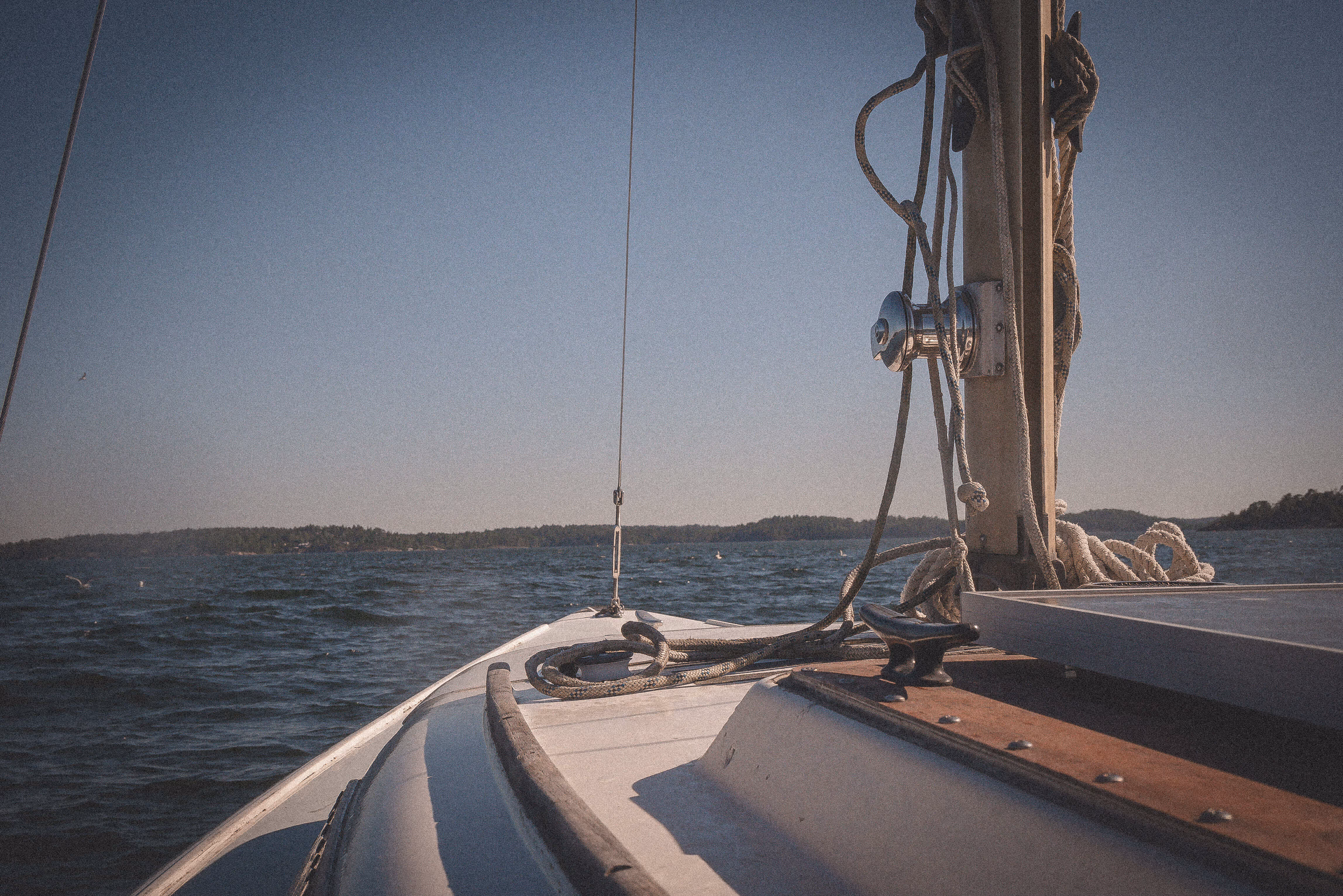 View from the bow of a sailboat on calm water with ropes and rigging against a clear sky and distant shoreline.