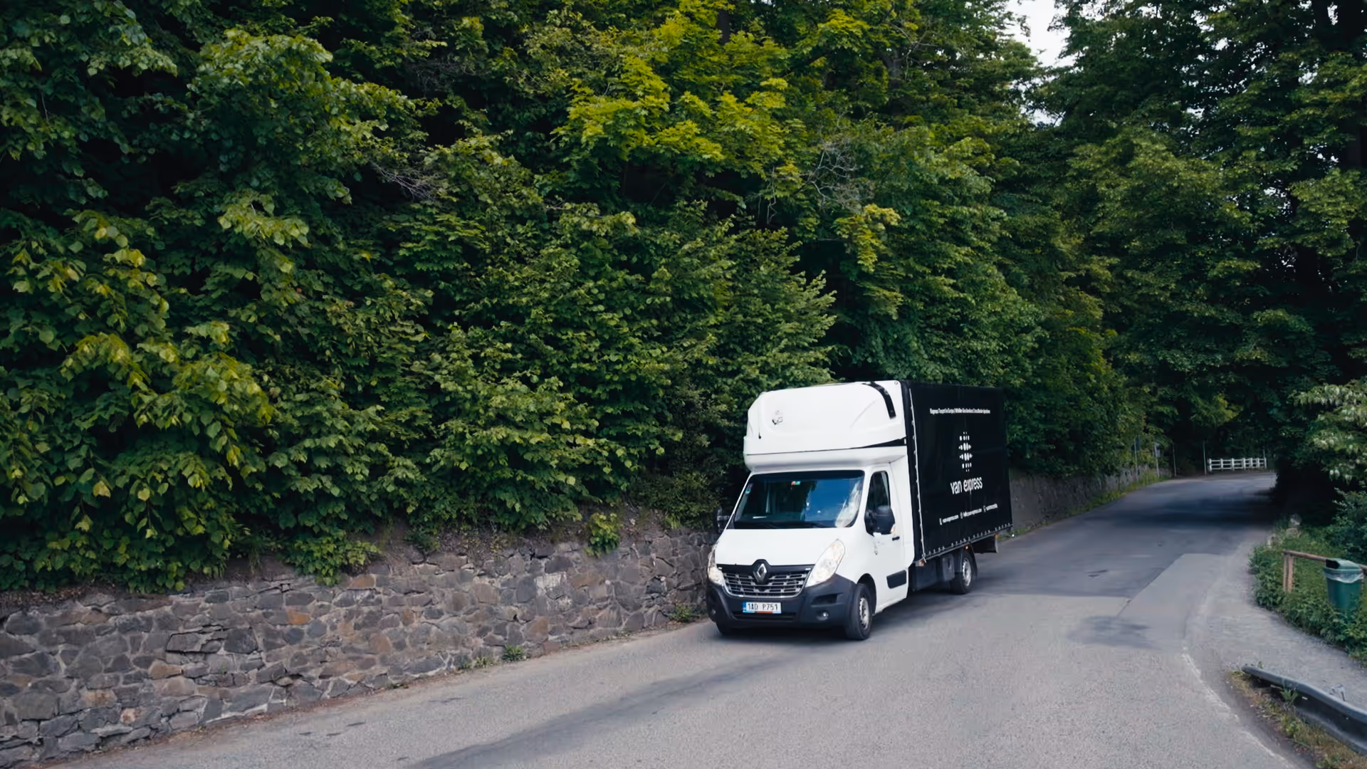 White delivery van with black Van Express branding driving on a road beside a stone wall and dense green trees.