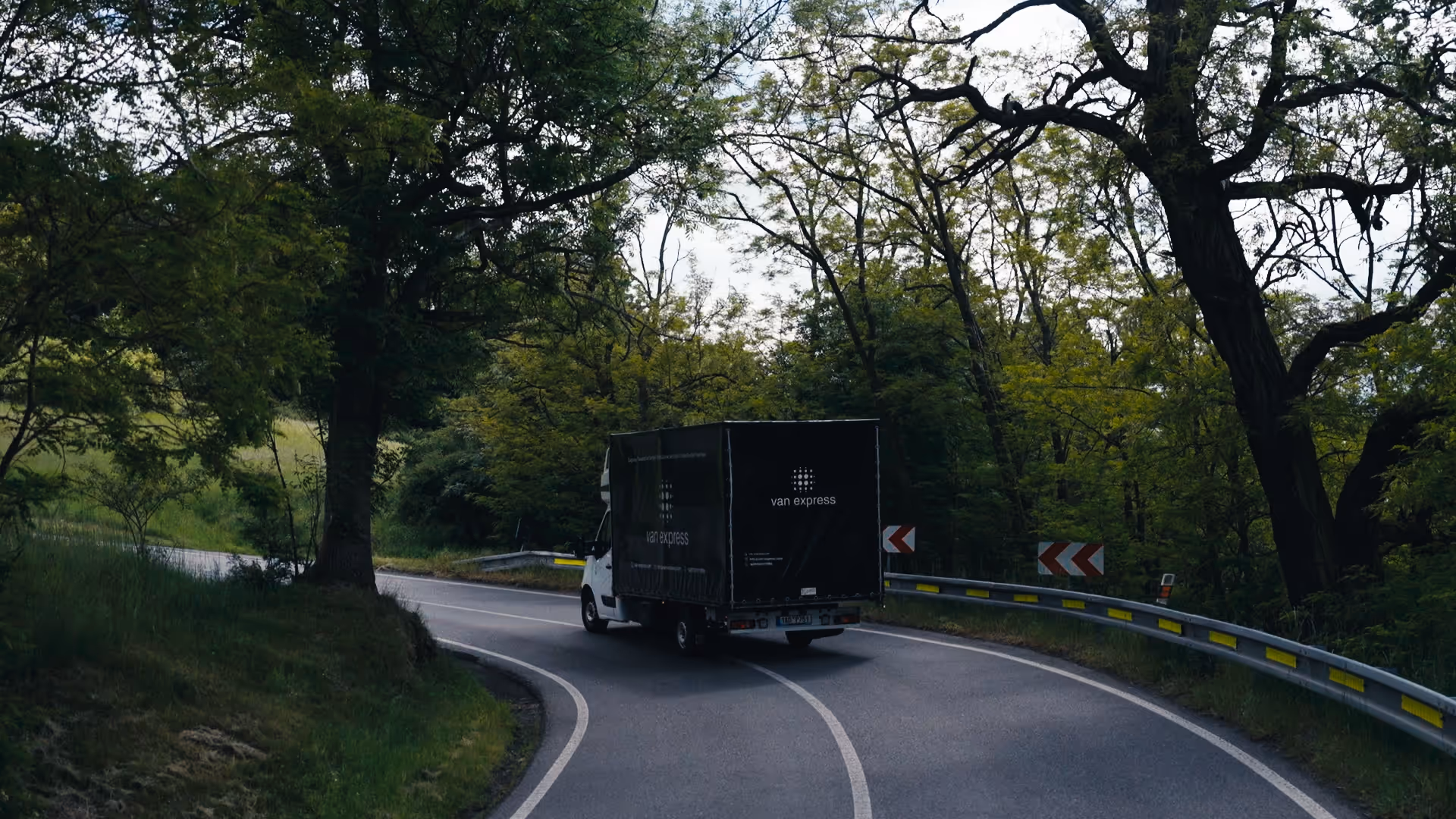 A white delivery truck with a black cargo cover labeled 'van express' driving on a winding road surrounded by green trees.