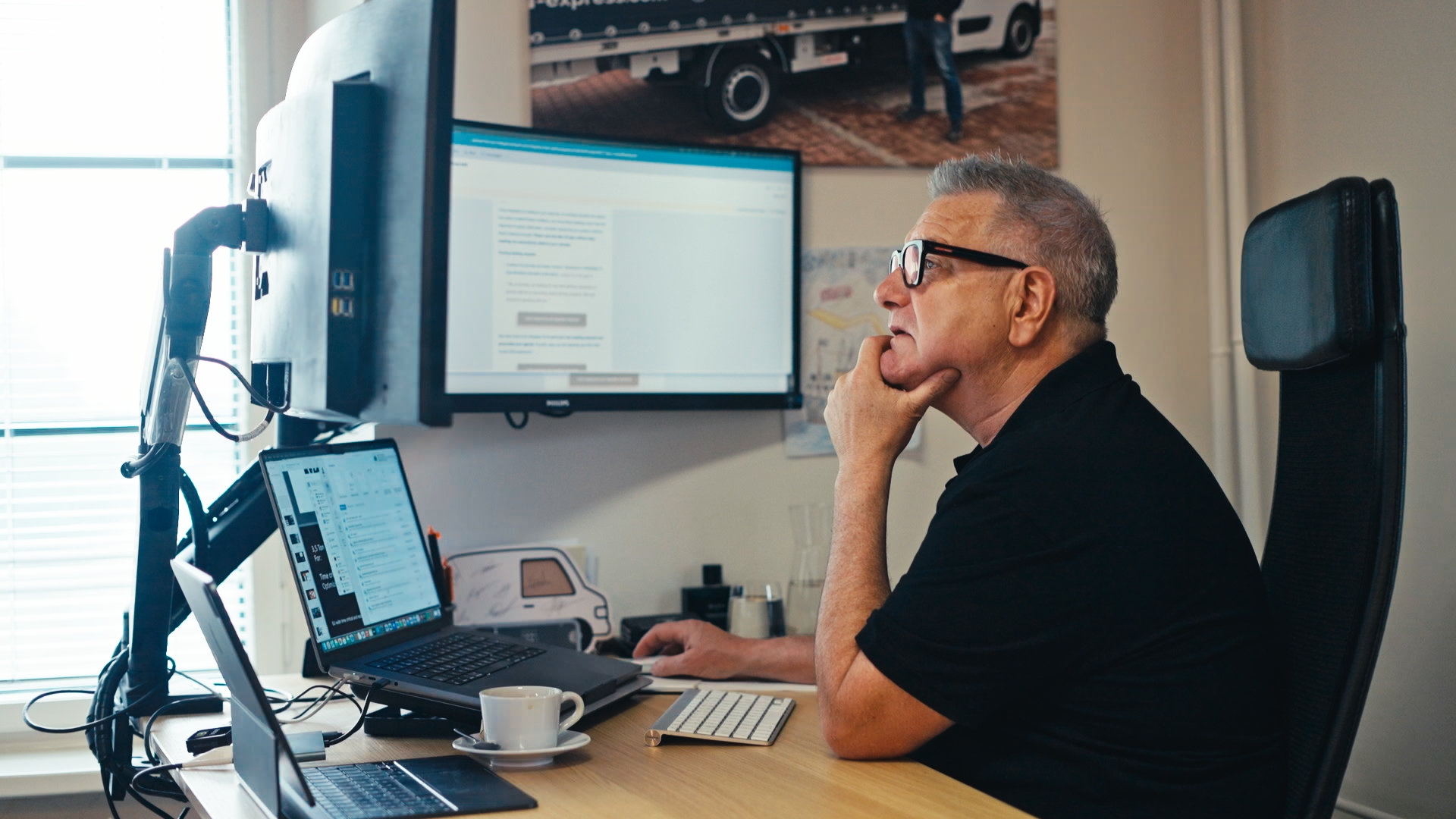 Older man with glasses sitting at a desk, looking thoughtfully at a large monitor while using a computer mouse.