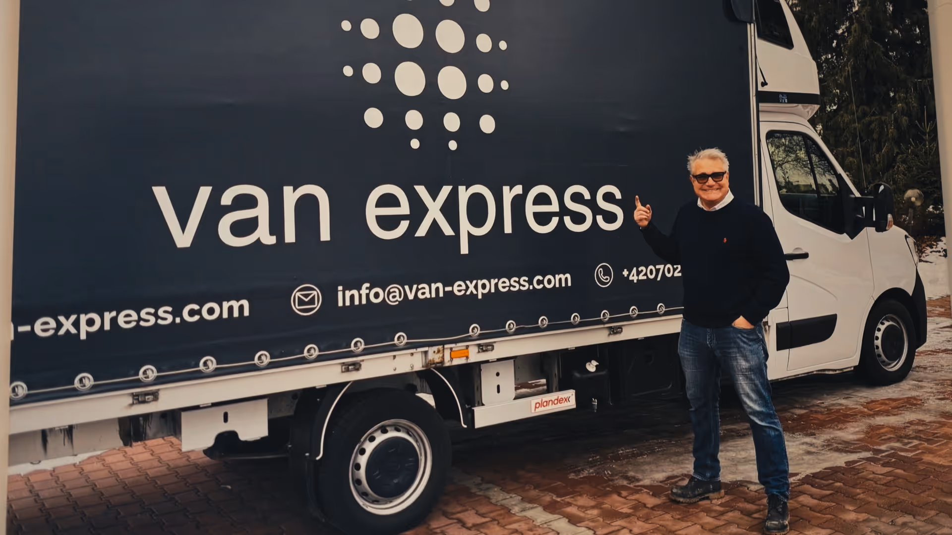 Smiling man with sunglasses pointing at a white van with a blue cover displaying 'van express' and contact info.