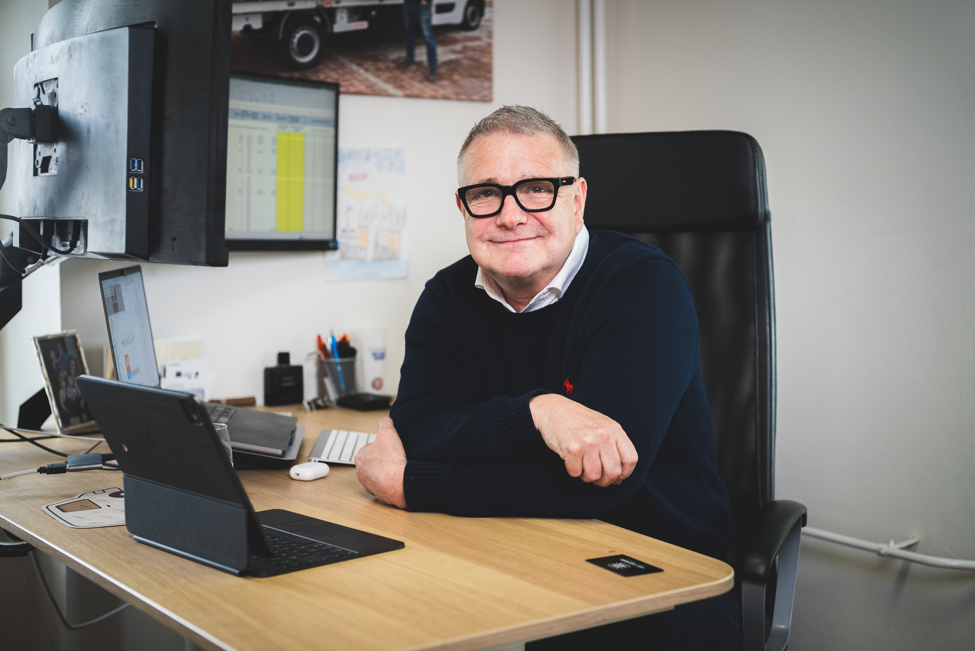 Smiling man with glasses sitting at a desk with a laptop, keyboard, and dual monitors in an office.