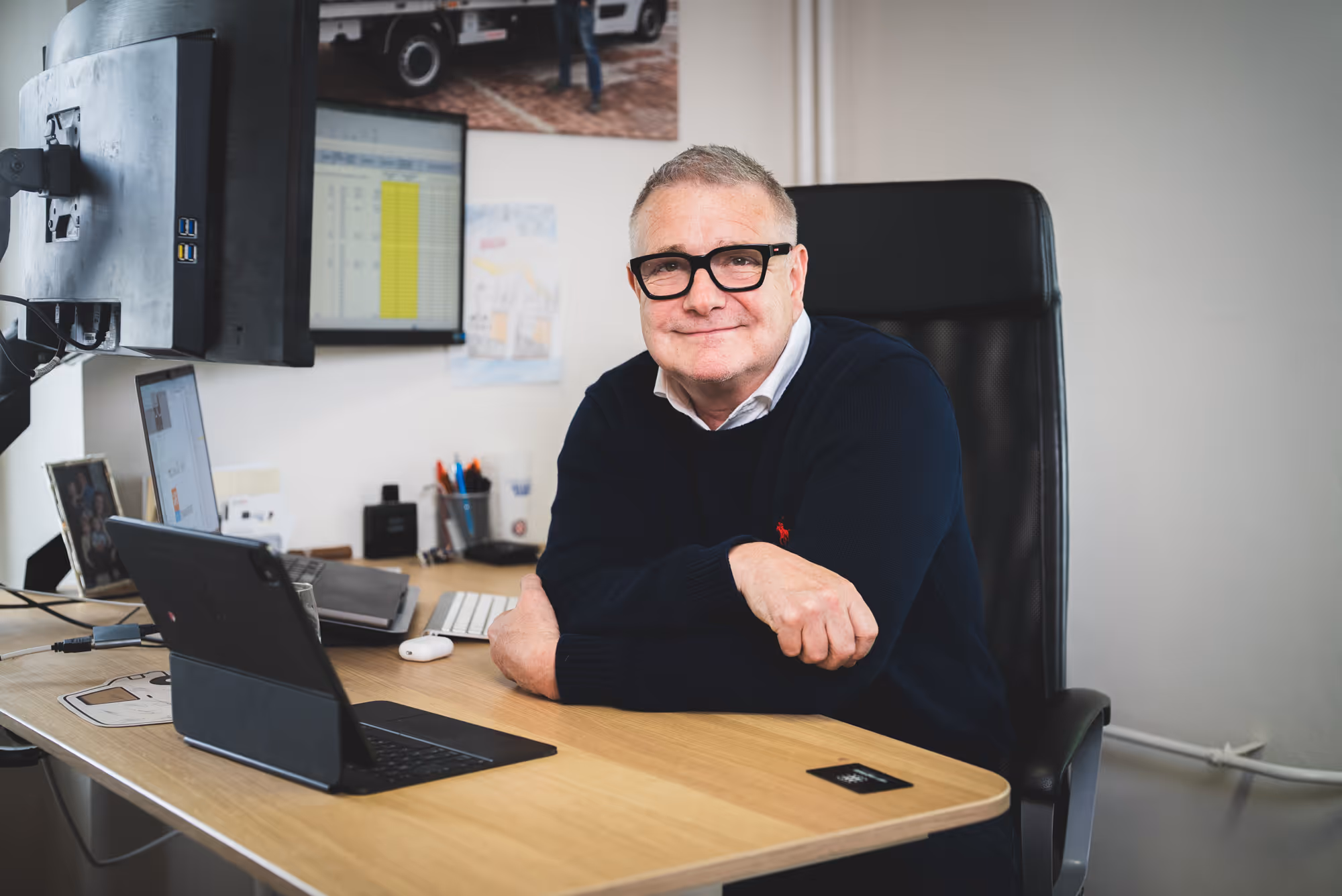 Smiling man with glasses sitting at a desk with a laptop, keyboard, and dual monitors in an office.