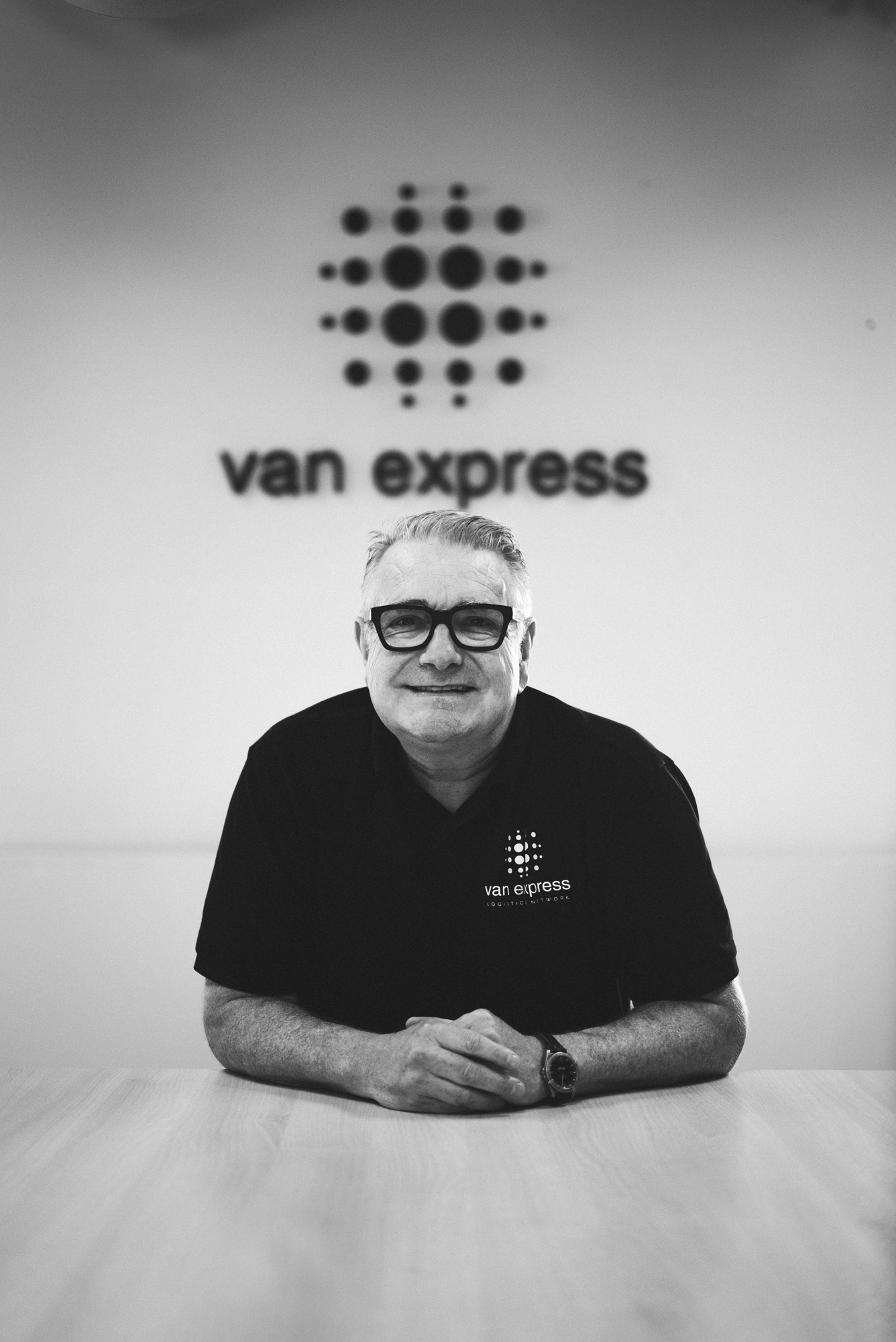 Smiling man with glasses sitting at a table wearing a black Van Express shirt with the company logo on the wall behind him.