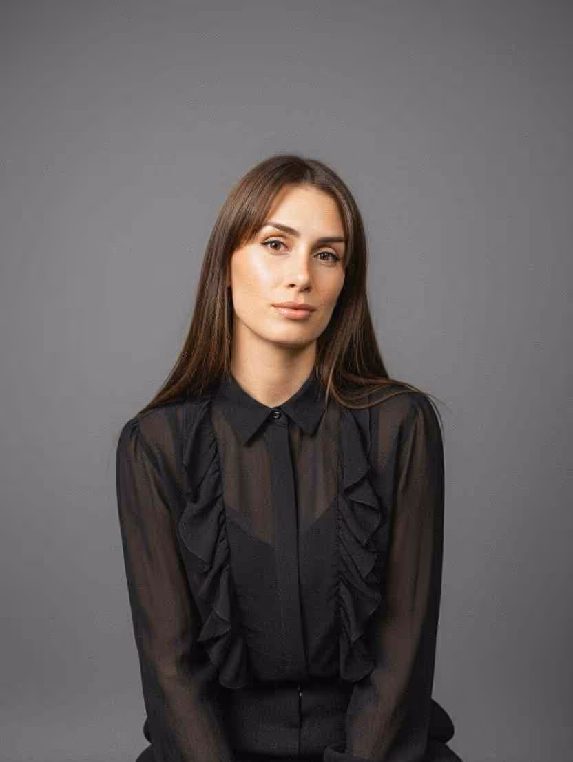 Portrait of a woman with long brown hair wearing a sheer black blouse with ruffles, against a gray background.