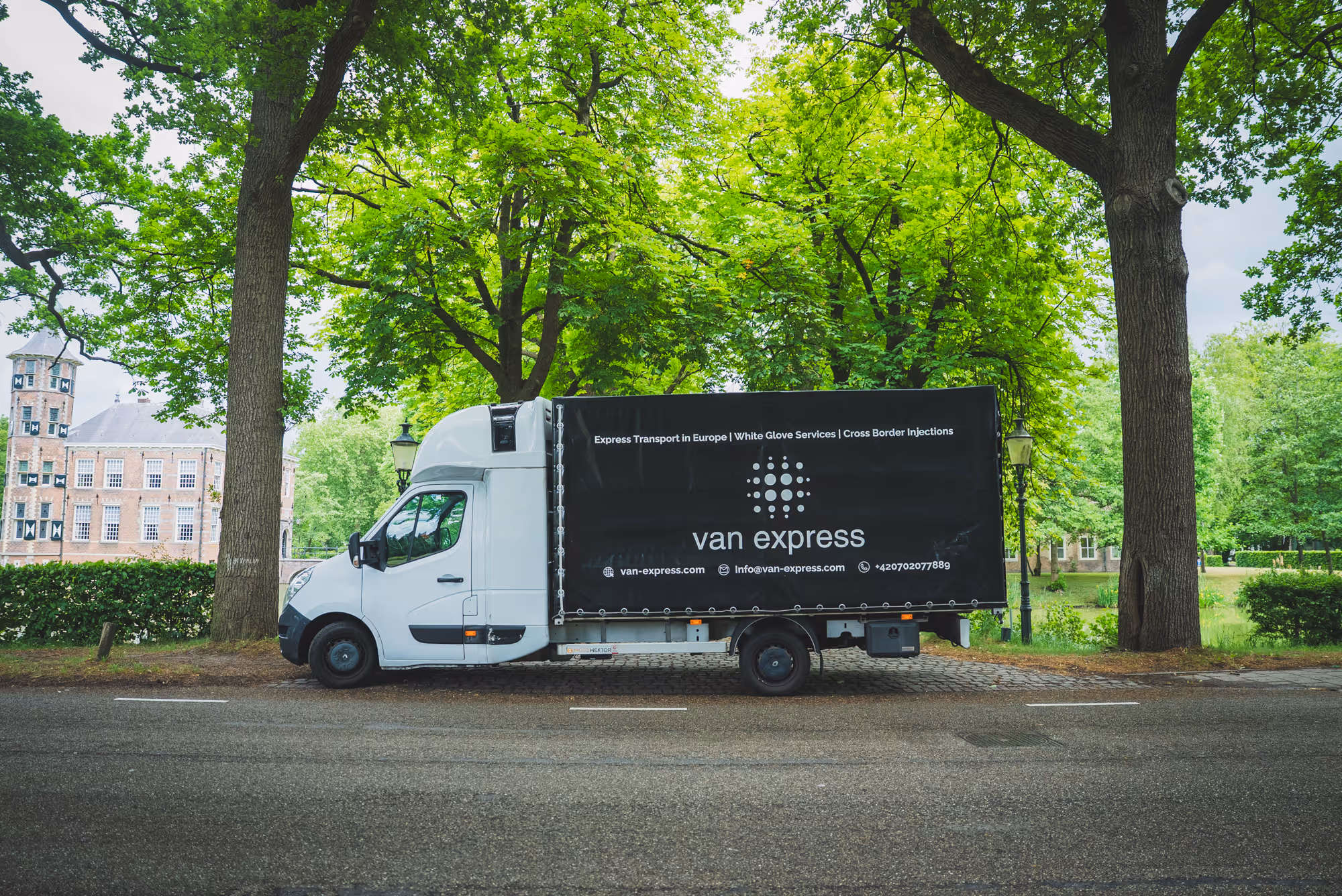 White van with black tarp reading 'van express' parked on a street lined with green trees and a historic brick building in the background.