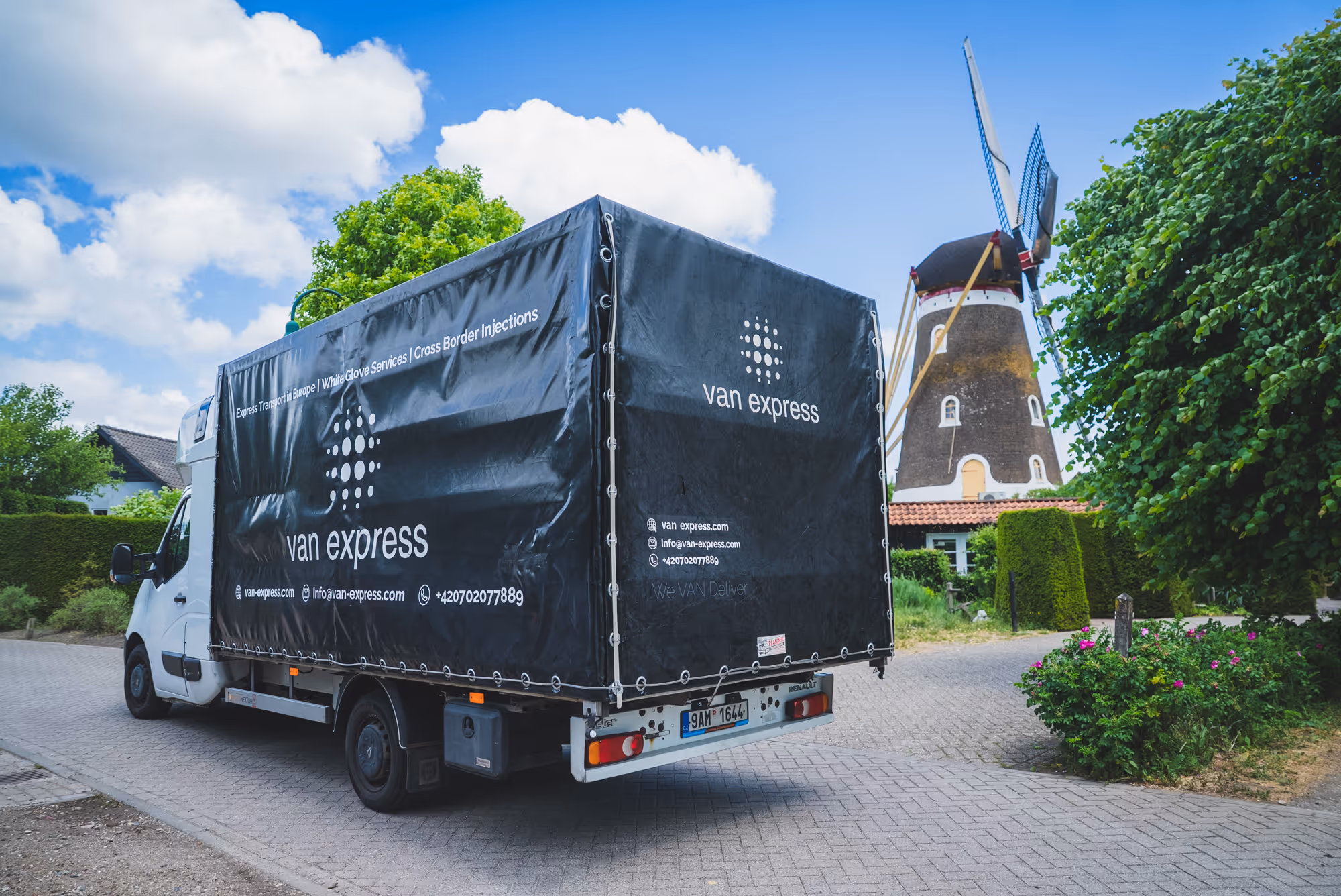 White delivery truck with black cover labeled 'van express' parked on a brick road near a traditional windmill and green foliage.
