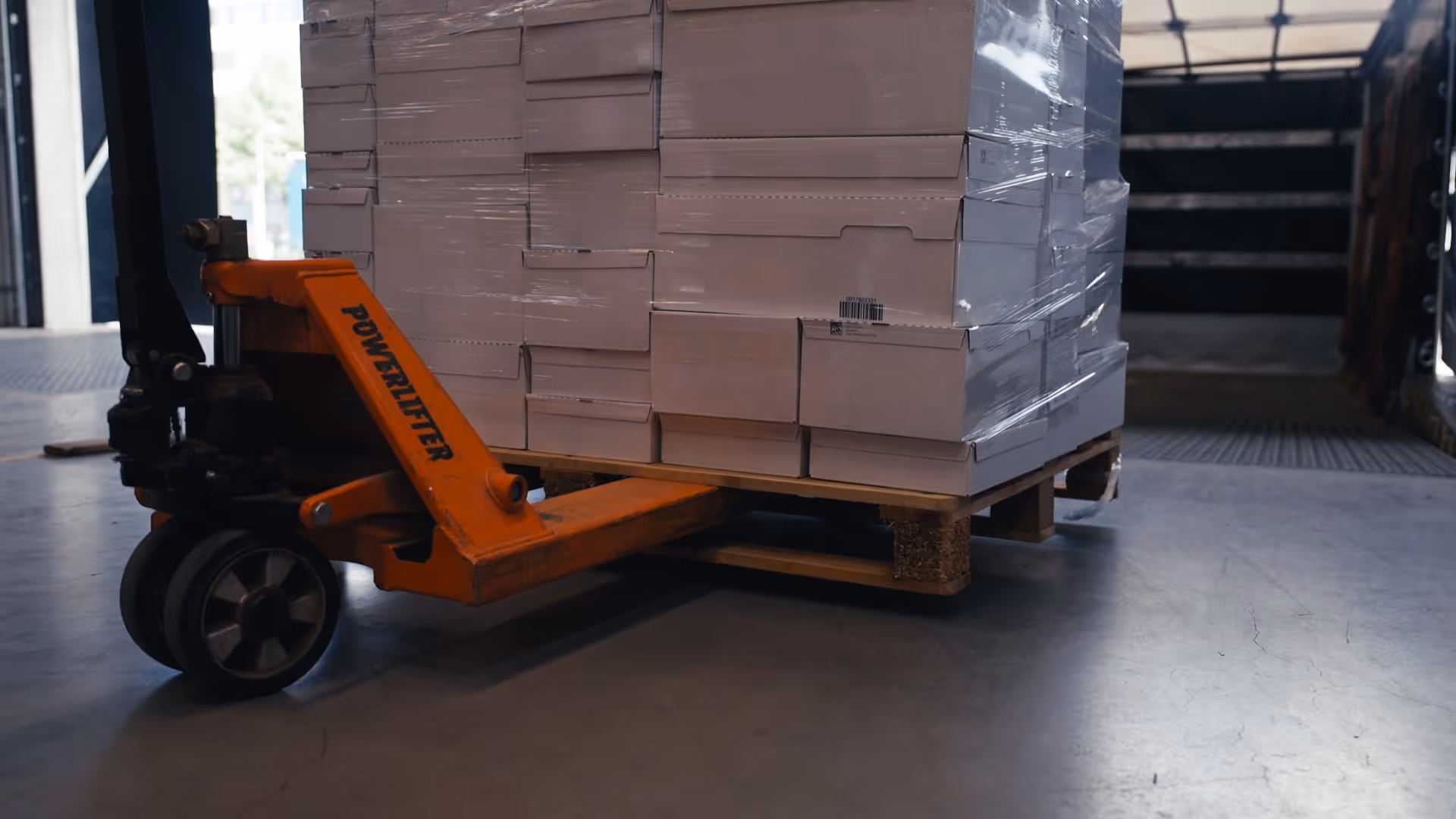 Orange Powerlifter pallet jack moving a wooden pallet stacked with wrapped cardboard boxes in a warehouse.